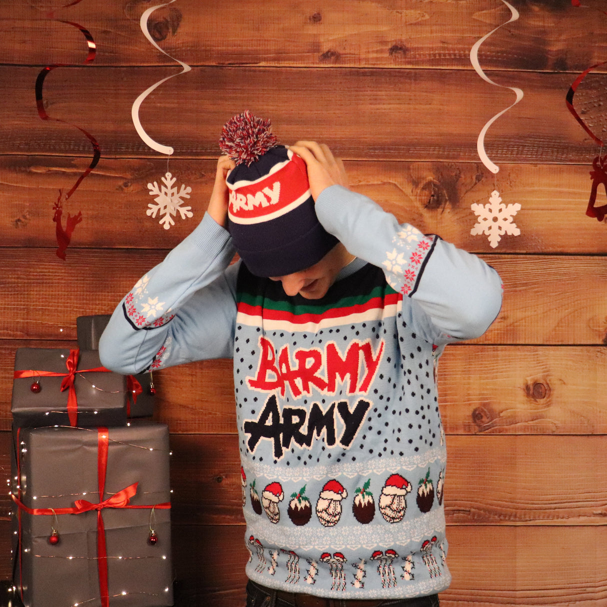 Person wearing a festive sweater with 'Barmy Army' text, standing against a wooden wall with Christmas decorations.